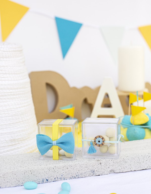 Μπομπονιέρες με διάφανα plexi κουτάκια διακοσμημένες με υφασμάτινο φιόγκο, ξύλινο στοιχείο μάτι και σατέν κορδέλα