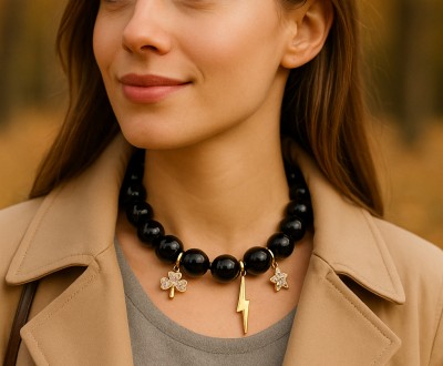 Κολιέ με μεγάλες μαύρες χάντρες και χρυσά μεταλλικά στοιχεία – τριφύλλι, αστραπή και αστέρι. Η αντίθεση μαύρου και χρυσού αναδεικνύει τη μοντέρνα κομψότητα και χαρίζει δυναμισμό σε κάθε look